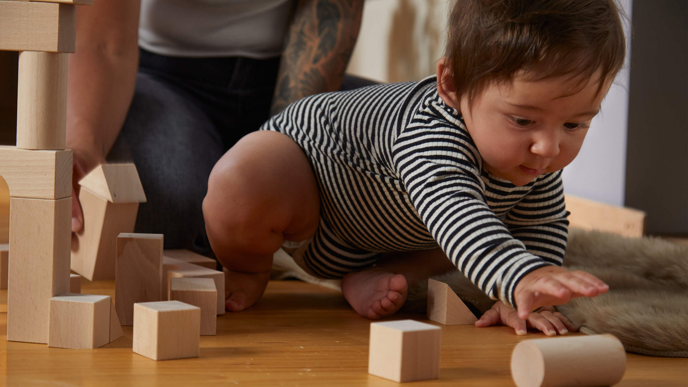 Toddle in a striped onesie sitting on the floor reaching for a cylinder shaped wooden block