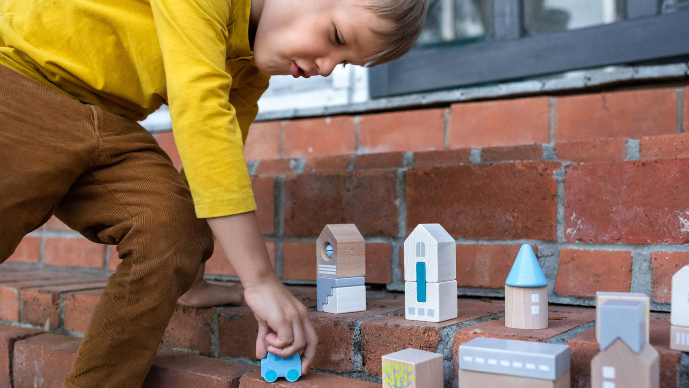 Boy outside on brick stairs moving a wooden car through building blocks set up like a town