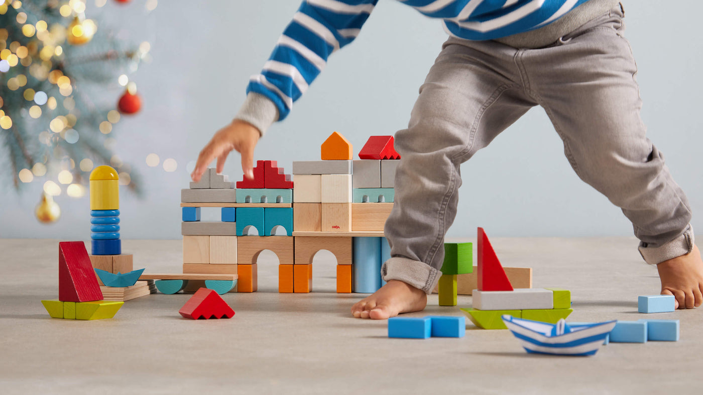 A child builds with colorful wooden blocks on a light floor, featuring a festive background with Christmas decorations.