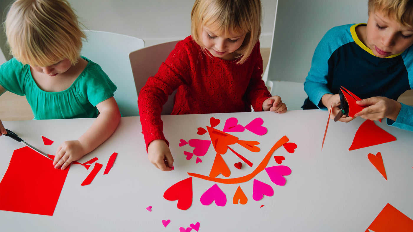 Overhead perspective of three kids sitting at a table cutting out paper hearts