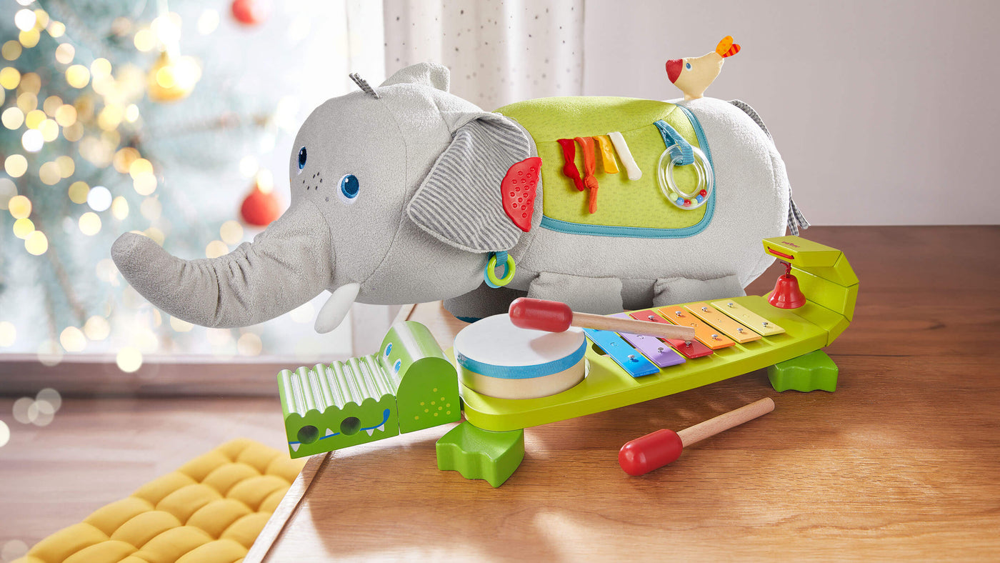 Plush elephant with ribbons and rings, and wood crocodile shaped xylophone with built in drum and bell on a wooden table and out of focus twinkle lights in the background