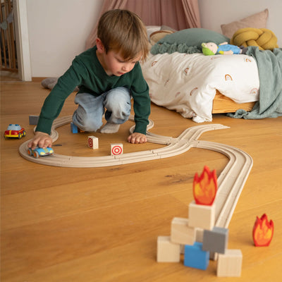 Child playing with a wooden car track on a wooden floor, with a bed and toys in the background.