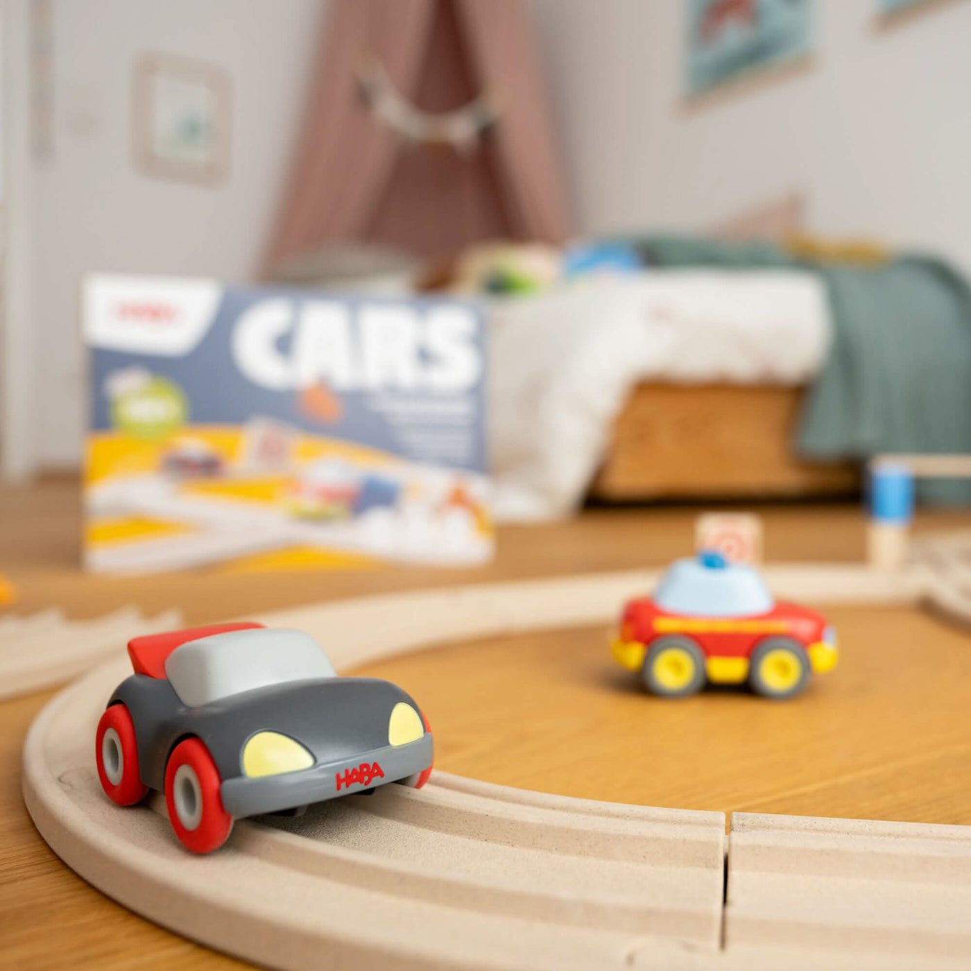 Toy cars on a wooden track in a child's bedroom with a blurred bed and a boxed set in the background.