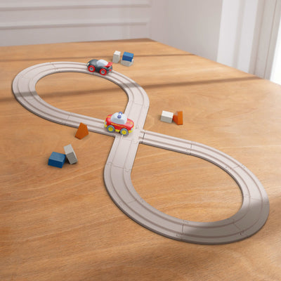 A toy racetrack in a figure-eight shape on a wooden table with two toy cars and various colored blocks.