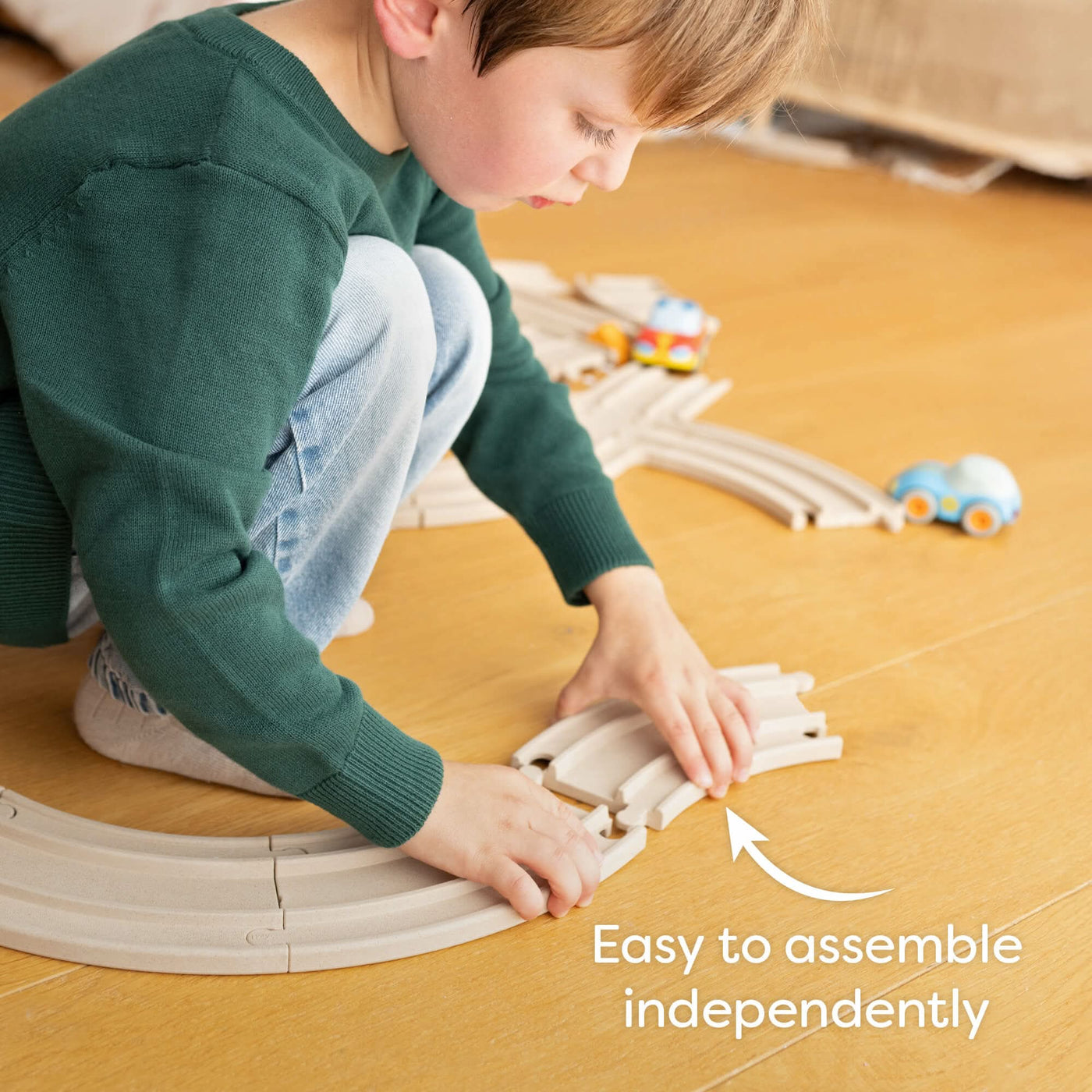 Child assembling a wooden track set on a floor with toy cars with the words "Easy to assemble independently."