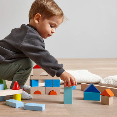 Young boy bending over and grabbing an orange wooden building block with other wooden blocks stacked in the background