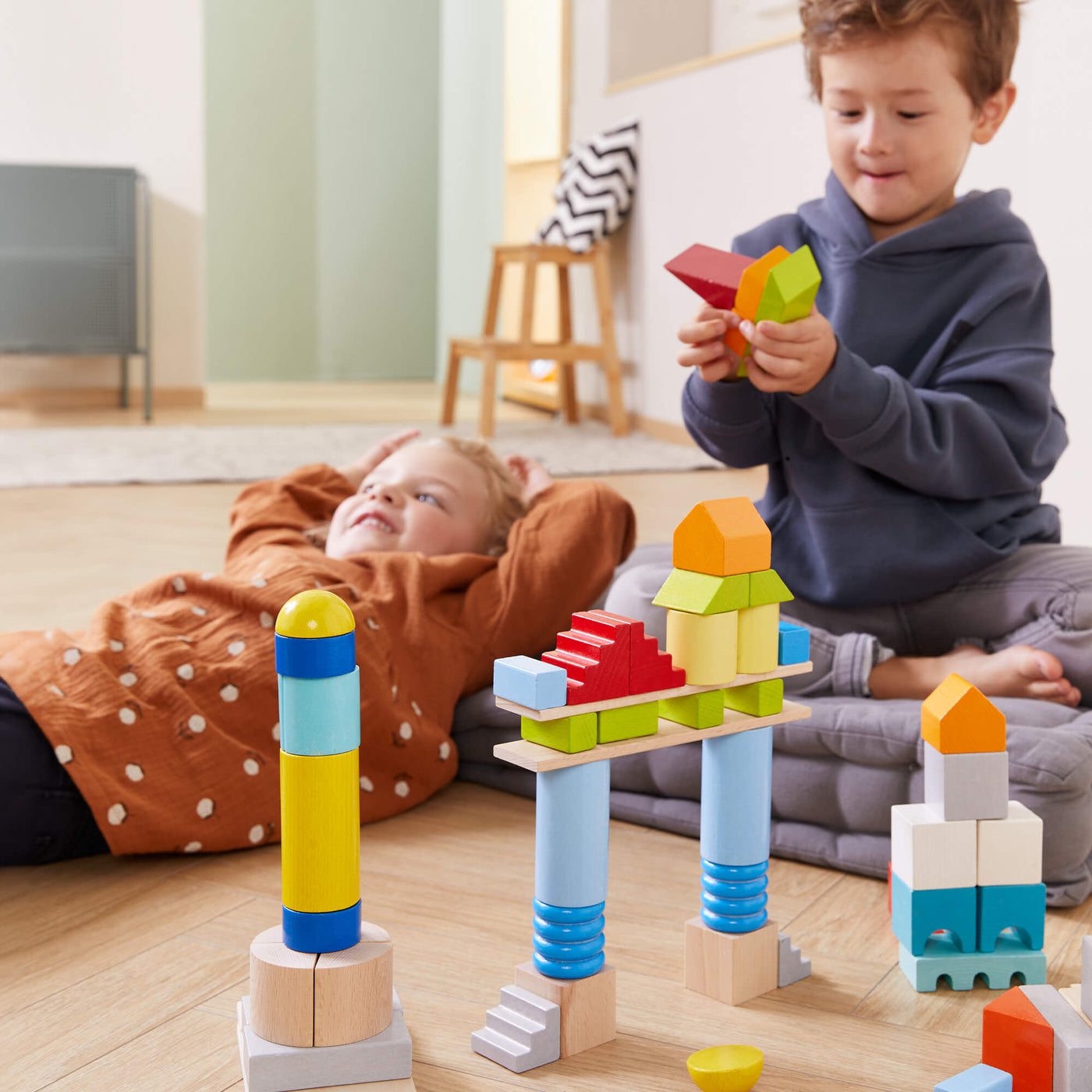 Two children play with colorful building blocks on a mat. One child constructs a tower, while the other lies down, smiling.