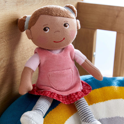 Snug Up Camila plush doll wearing a pink dress with striped leggings sitting on a colorful rainbow shaped cushion.