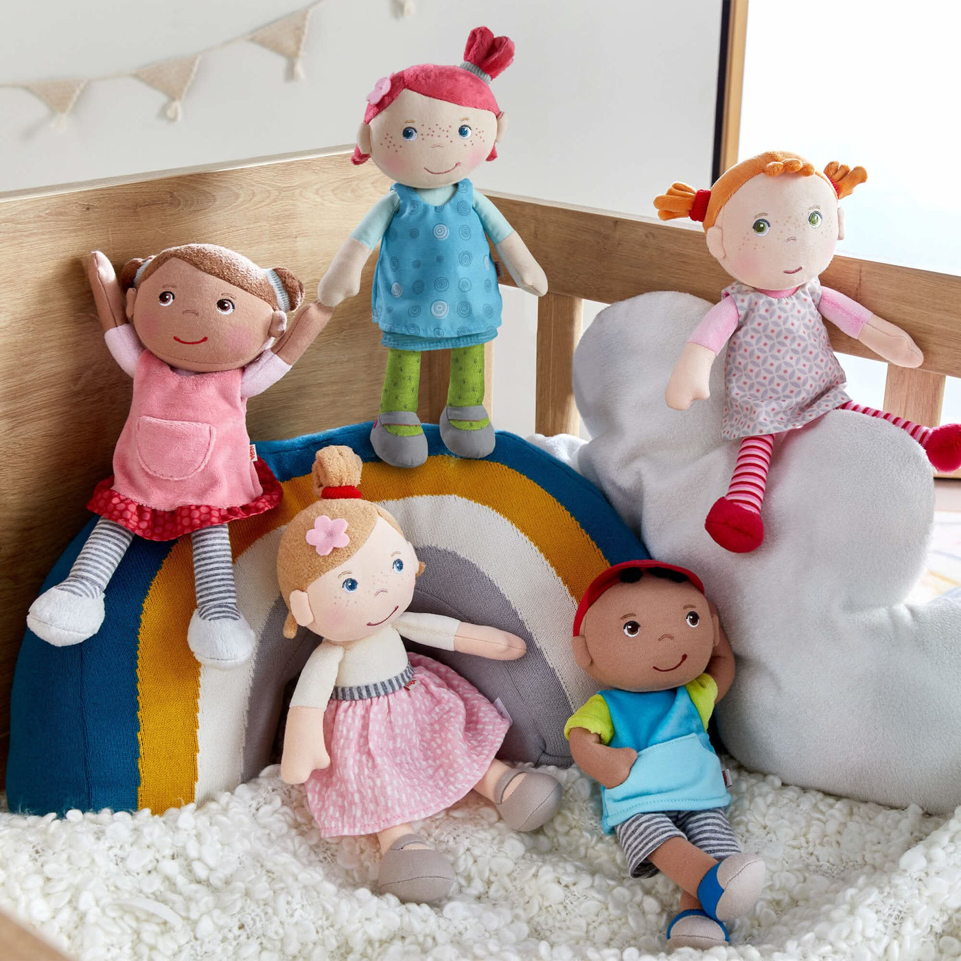 Five plush dolls posed inside a wooden crib with rainbow and cloud cushions.