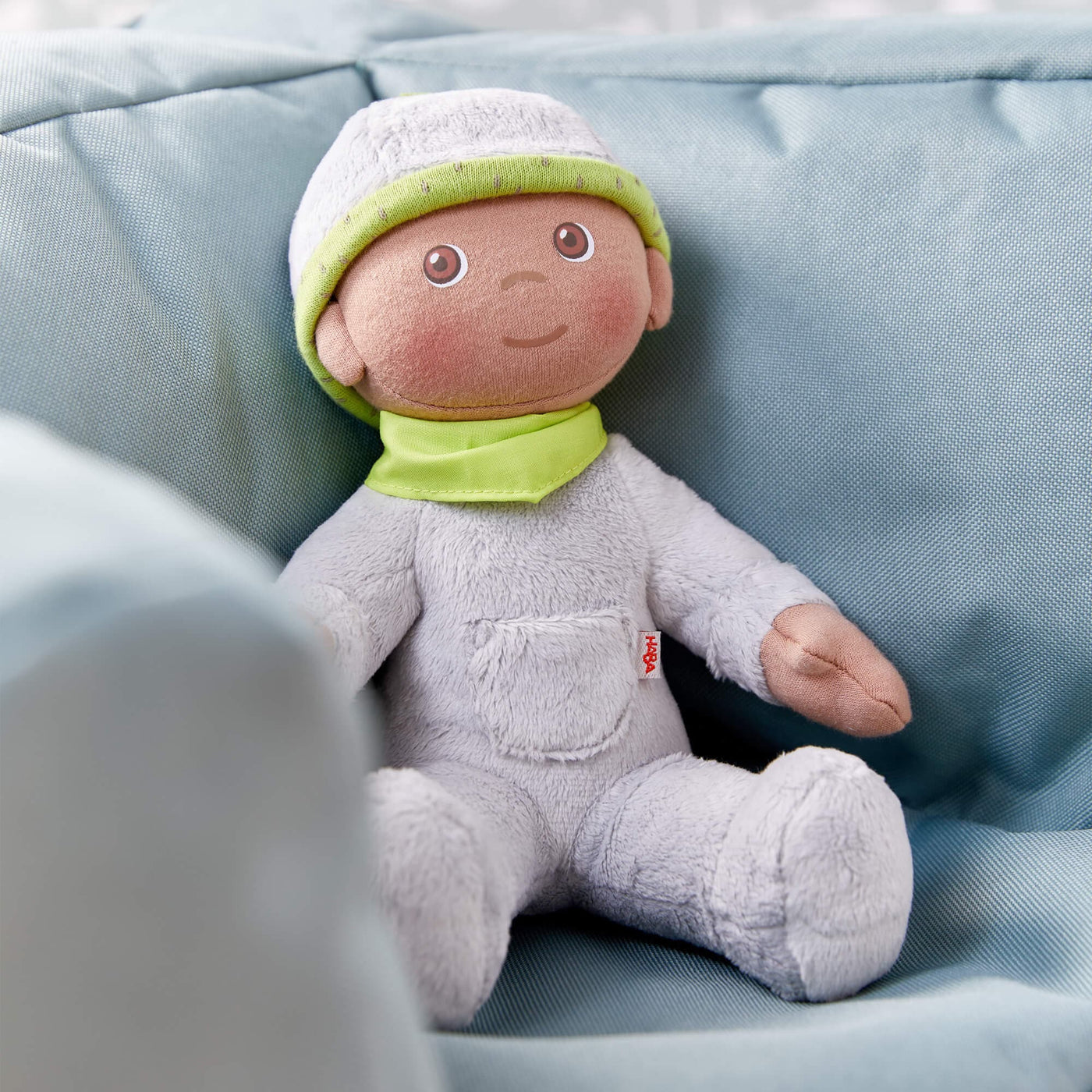 HABA Snug Up Doll Ollie with grey fleece body, green bandana, printed face, and grey fleece cap with green band sitting on a couch