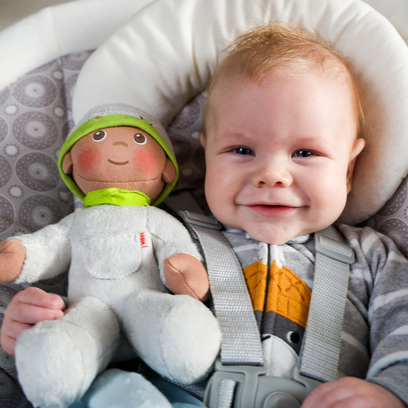 A smiling baby in a carrier with a plush doll wearing a gray and green hat.