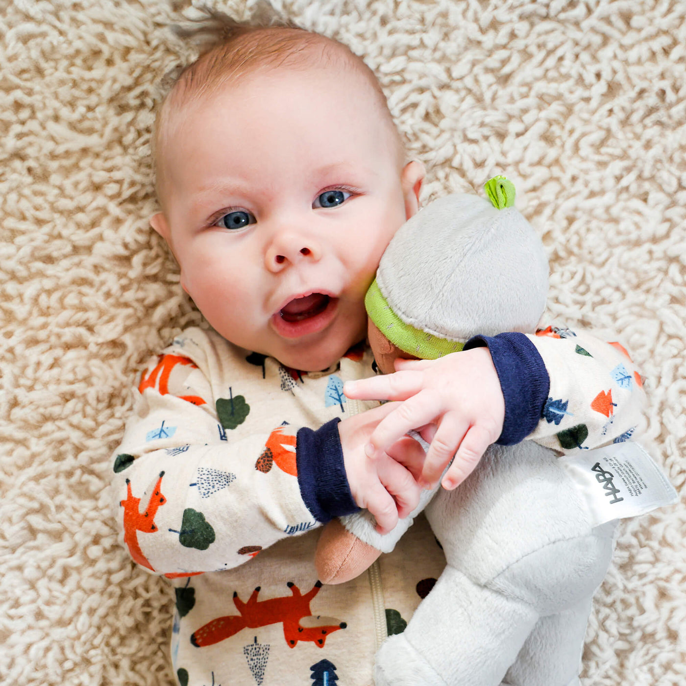 Baby in a patterned onesie holding a gray plush doll on a fluffy rug.