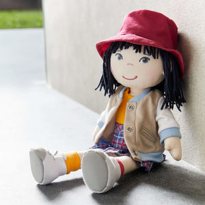 Yuki cloth doll wearing a red hat, tan jacket, yellow shirt, and checkered skirt sits against a wall.