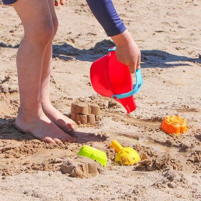 A child in a blue shirt stands barefoot on the sand, pouring water from a red bucket onto colorful sand molds.