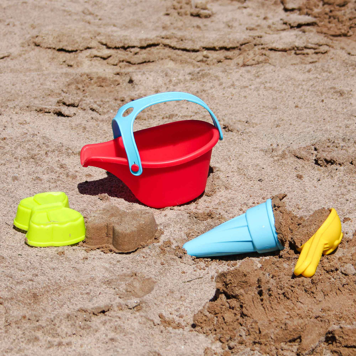 A red plastic bucket, blue cone-shaped mold, yellow shovel, and green sand molds sit on a sandy beach.