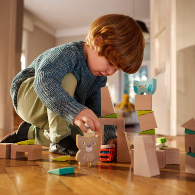 A young boy kneels on a hardwood floor, building a tower with wooden blocks, bear, and an owl.