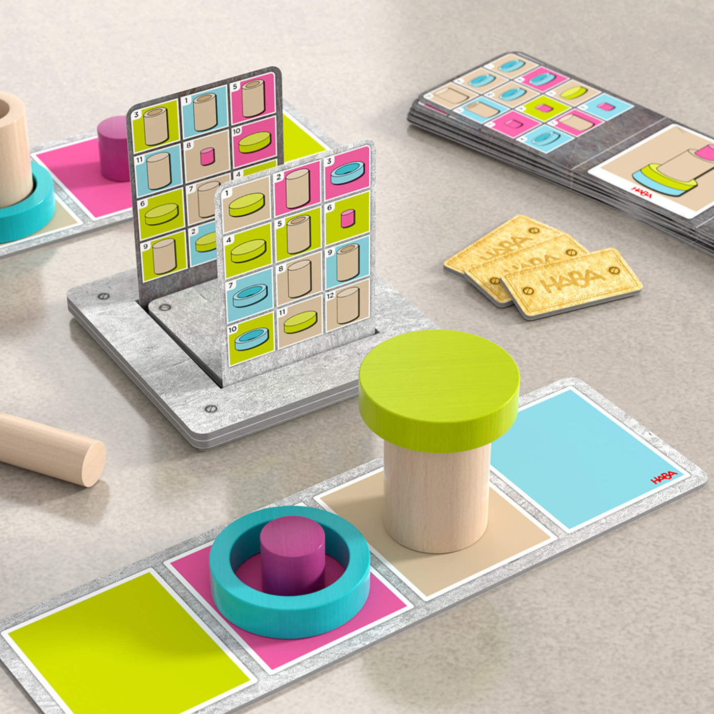 A tabletop board game involving colorful blocks and cards. In the foreground, there are two cylindrical wooden blocks on a game card with squares in green, pink, and blue. 