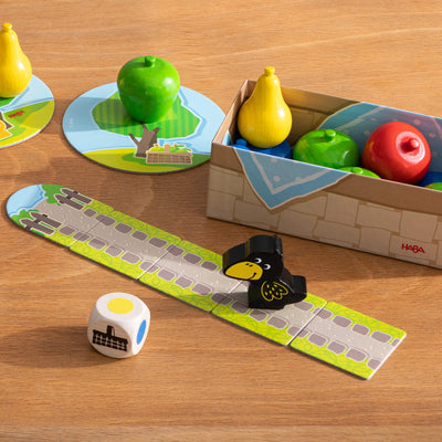 Colorful game pieces from the game First Orchard including fruit, a raven figure, dice, and a path on a wooden surface, with a gameplay box visible.