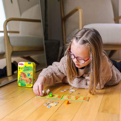 A young girl playing a board game on a wooden floor with the game box "Clippety-Clop!" nearby.