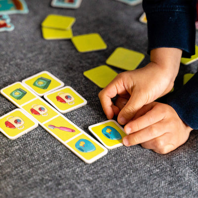 A child's hand places a colorful card on a pile of matching game cards arranged on a gray sofa.