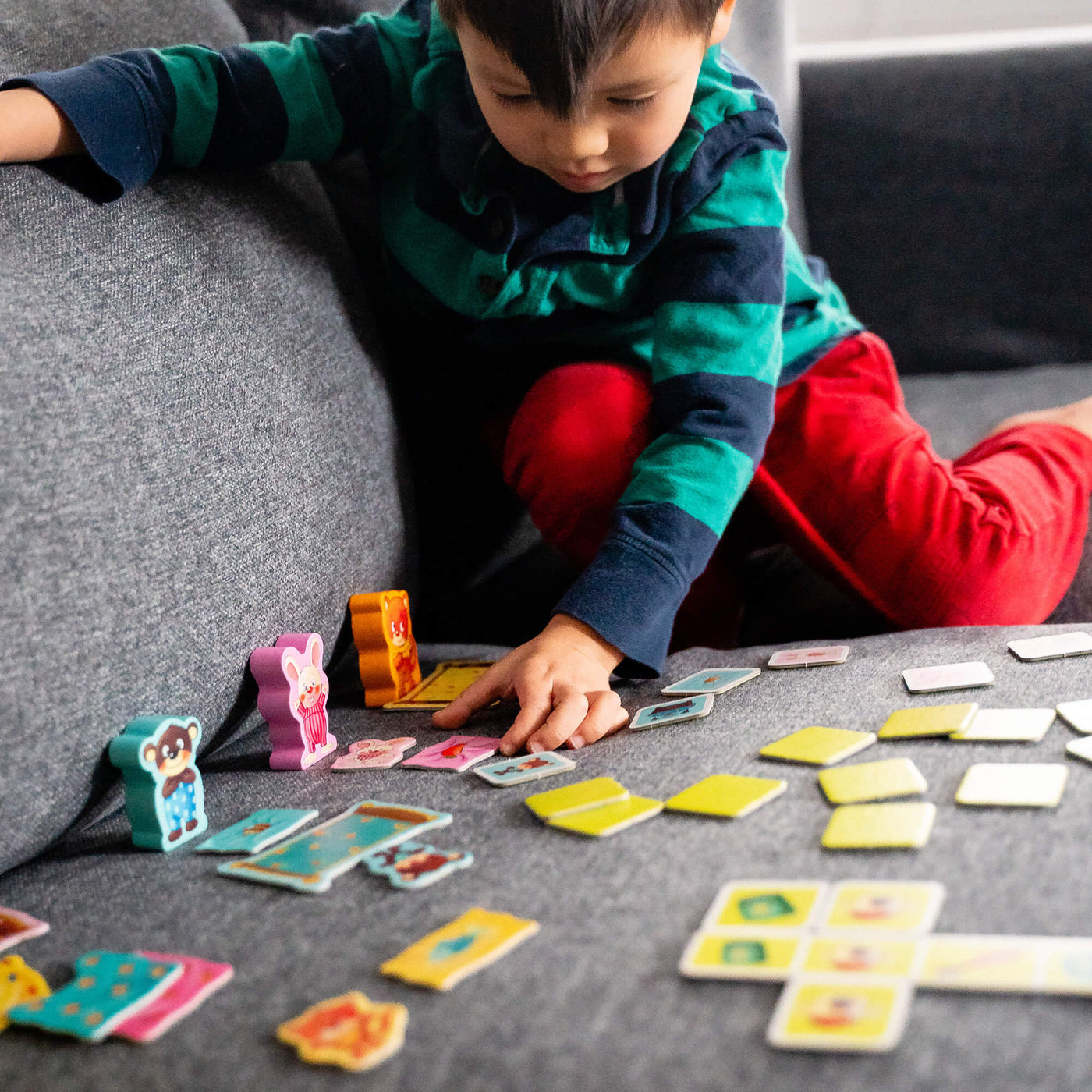 A young child in a striped shirt and red pants plays My Very First Games - Off To Bed! on a sofa, arranging colorful game pieces spread out around them.