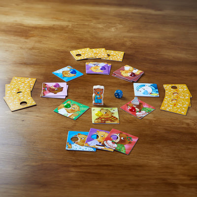 Cardboard bear game pieces with wooden party hat, blue die, green, yellow, red, light blue, blue, brown, purple, light purple, and green wooden noses, and wooden bear figure on a wooden table