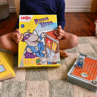 Child sitting on the floor holding a Giant Rhino Hero XXL game box with pieces and a card stack next to them