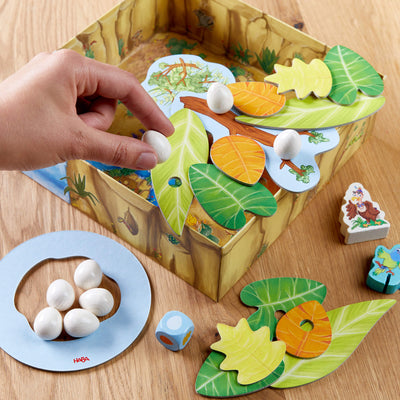 Close-up of a colorful board game with a hand placing a white egg piece onto illustrated leaf pieces in a cardboard box.