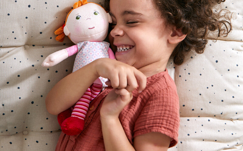 A child smiling while hugging a red-haired Roya doll on a speckled cushion.