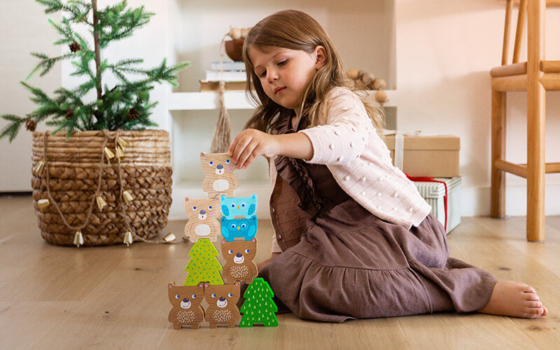 Girl wearing a dress and sweater sitting on the floor placing a wooden bear on a stack of wooden bears and trees with presents and an evergreen behind her