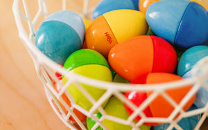 A white wire basket filled with colorful Fun with Sounds Wooden Eggs on a wooden surface.