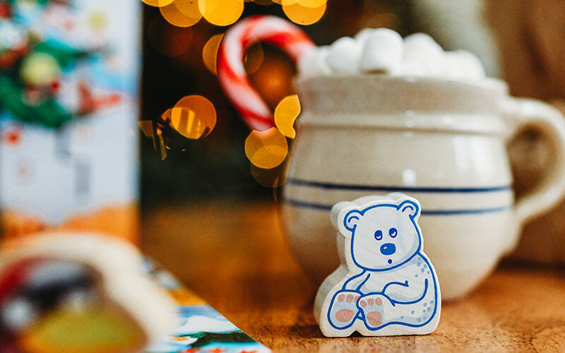 Wooden bear in front of a mug of hot coco with marshmallows and a candy cane in the background