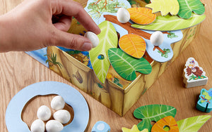 A hand holding a white egg game piece over a jungle-themed board game with colorful leaves.