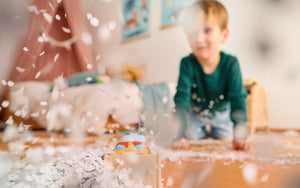 A child plays with confetti indoors, with a colorful toy HABA Car in the foreground.