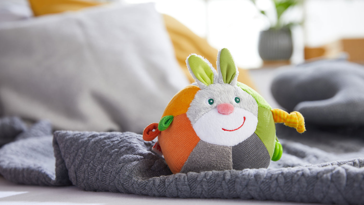 Colorful plush toy shaped like a ball with bunny ears and face on a gray blanket.