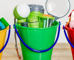 Three colorful buckets with various toys and objects, including a magnifying glass and wooden blocks, on a wooden surface.