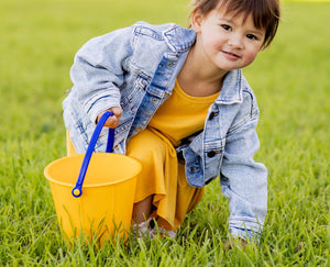 Child in a yellow dress and denim jacket holding a yellow Spielstabil bucket on a grassy field.