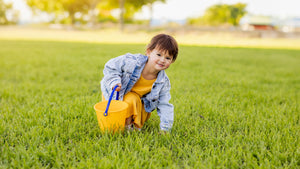 Child in a yellow dress and denim jacket holding a yellow Spielstabil bucket on a grassy field.