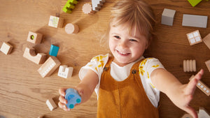 Child smiling and reaching toward the camera surrounded by wooden blocks and toys on the floor.