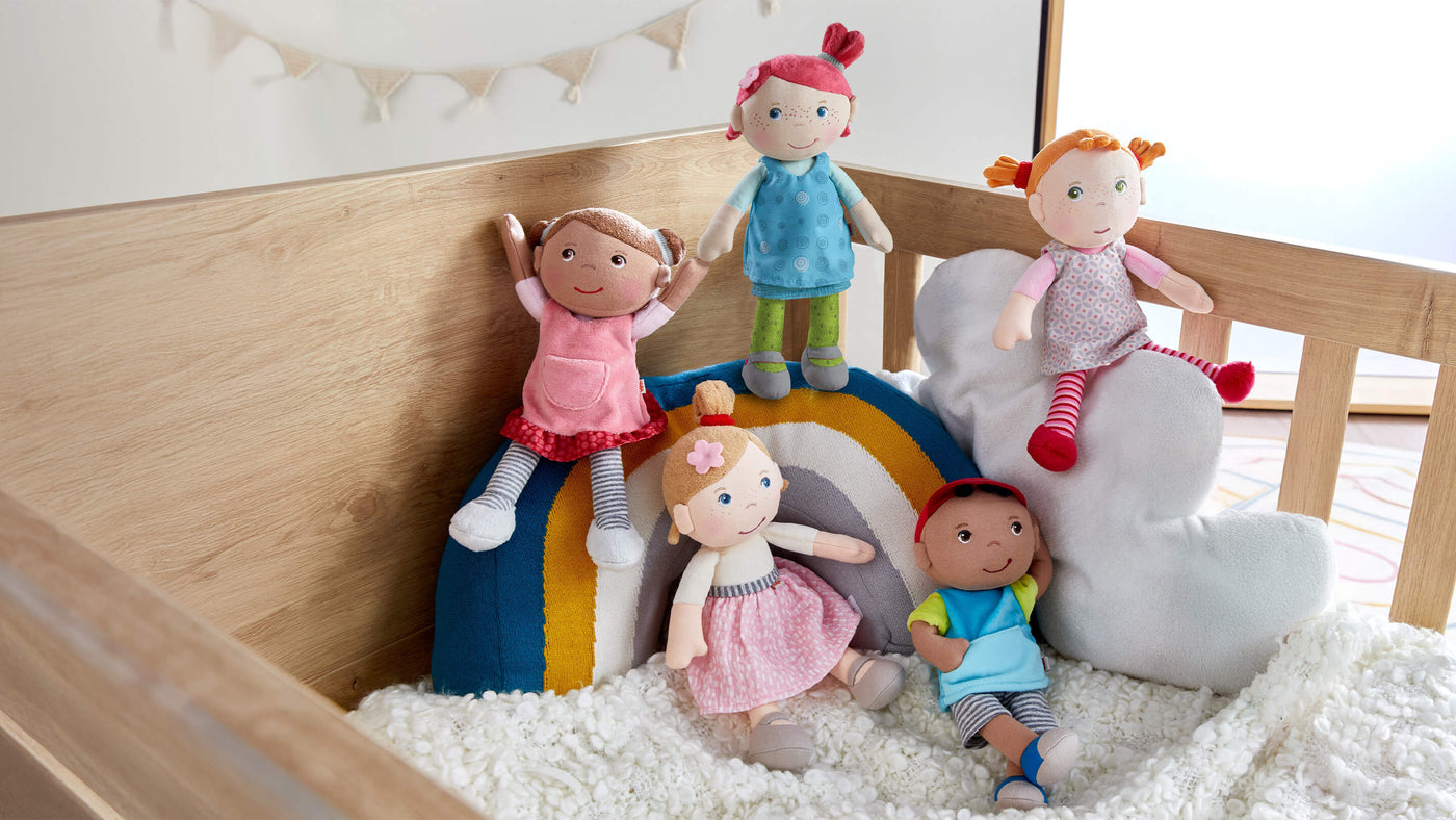 Five colorful fabric dolls arranged in a wooden crib with a fluffy blanket and rainbow cushion.