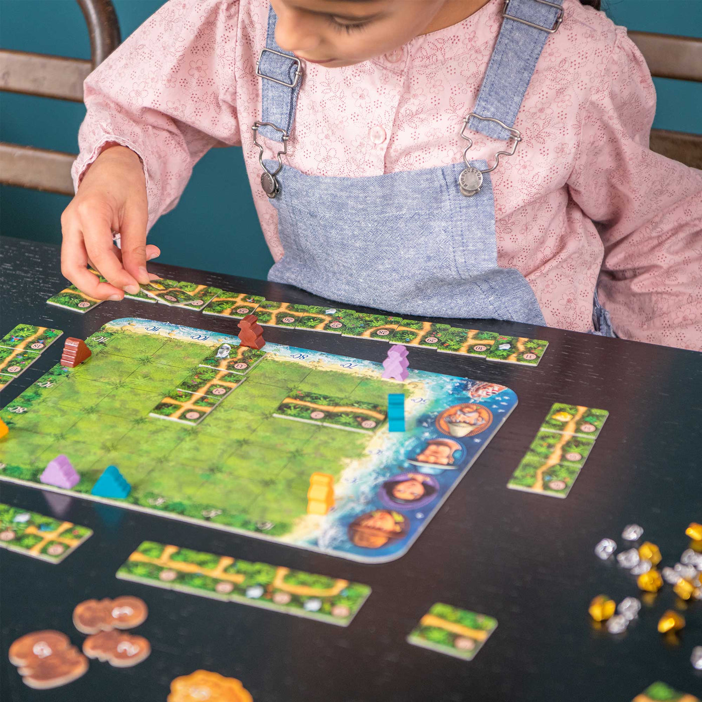 A child with a pink shirt and denim overalls plays Karuba board game, placing colorful pieces on a green game board.