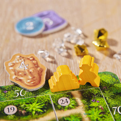 A close-up of Karuba board game featuring yellow player pieces, gems, and a token with a face, set on a lush, green game board.