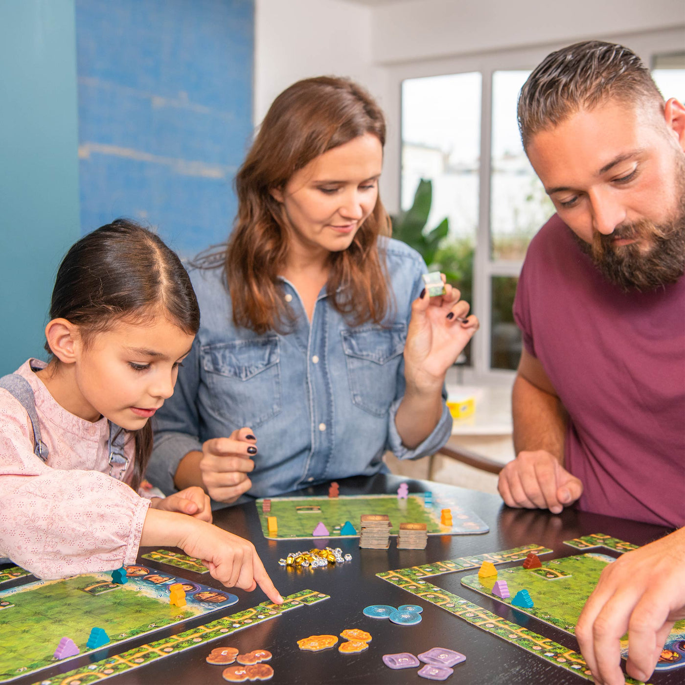 A young girl, a woman, and a man gather around a table with the Karuba board games, enthusiastically engaged in play.