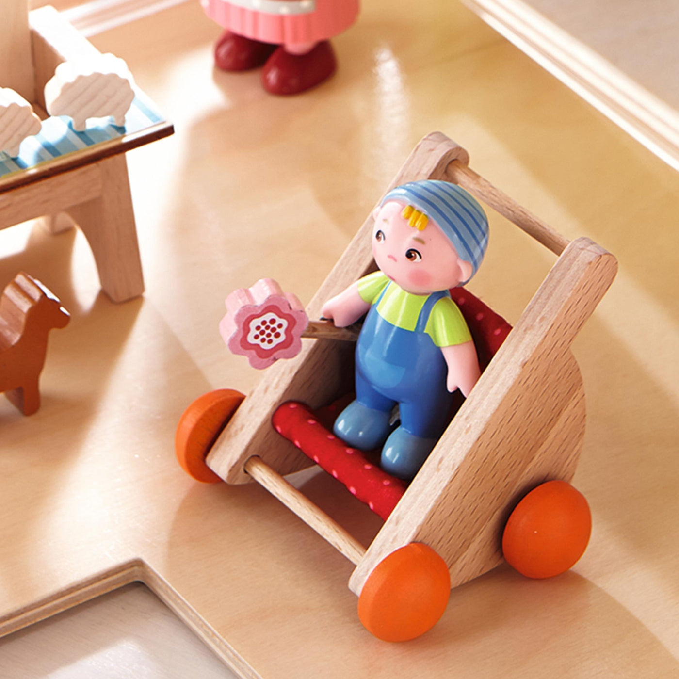 A doll in a wooden toy walker with orange wheels holding a pink flower toy, surrounded by wooden toys on a light surface.