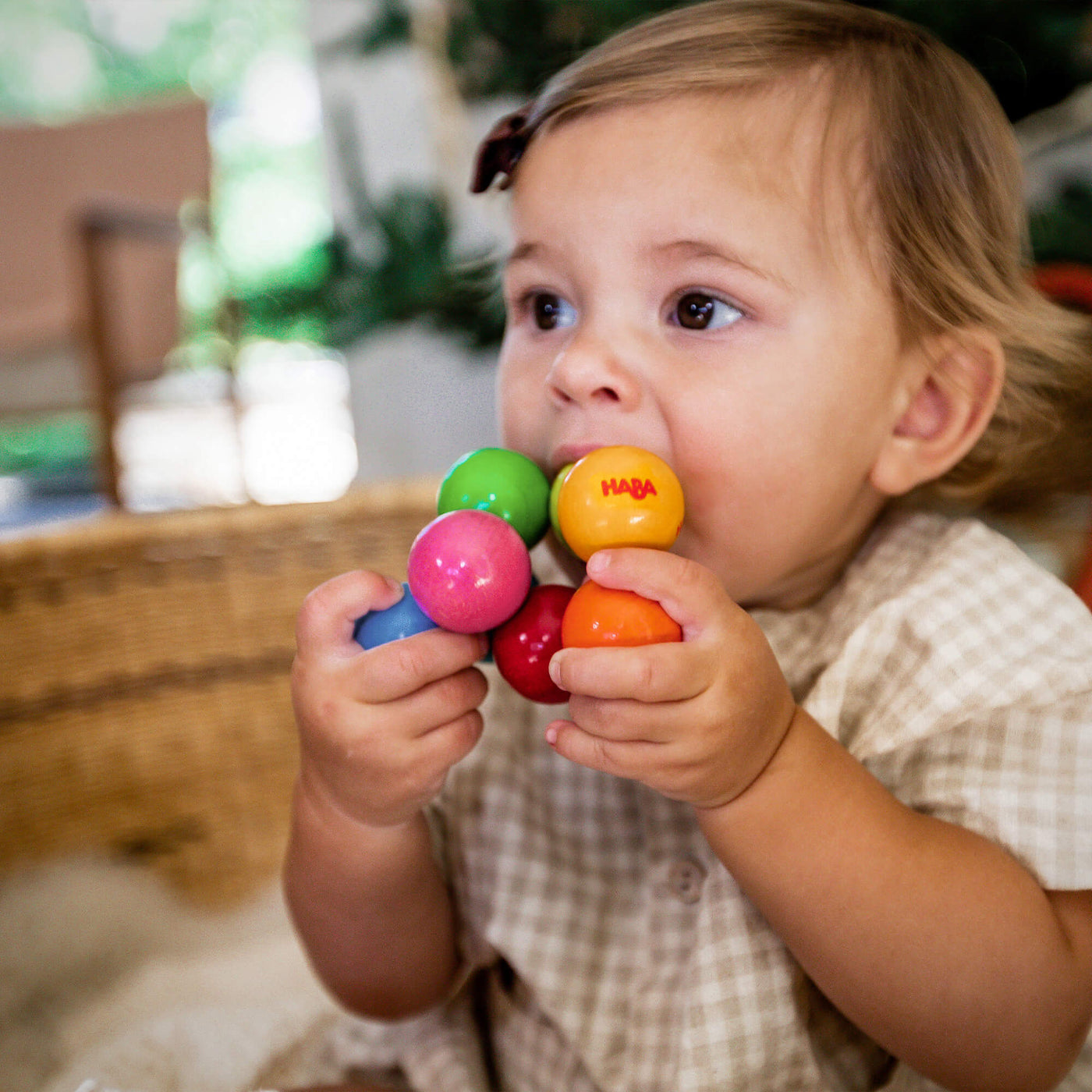 A toddler in a basket holds and chews on a Magica Wooden Clutching Toy made of colorful wooden balls.