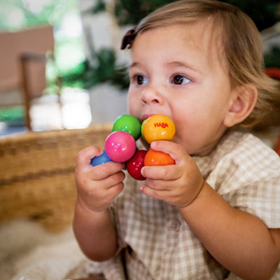 A toddler in a basket holds and chews on a Magica Wooden Clutching Toy made of colorful wooden balls.