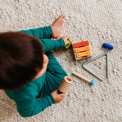 A young child sitting barefoot on a textured carpet holding various musical toys. Near the child is a small wooden xylophone, a triangle percussion instrument with a blue handle and a separate wooden stick.