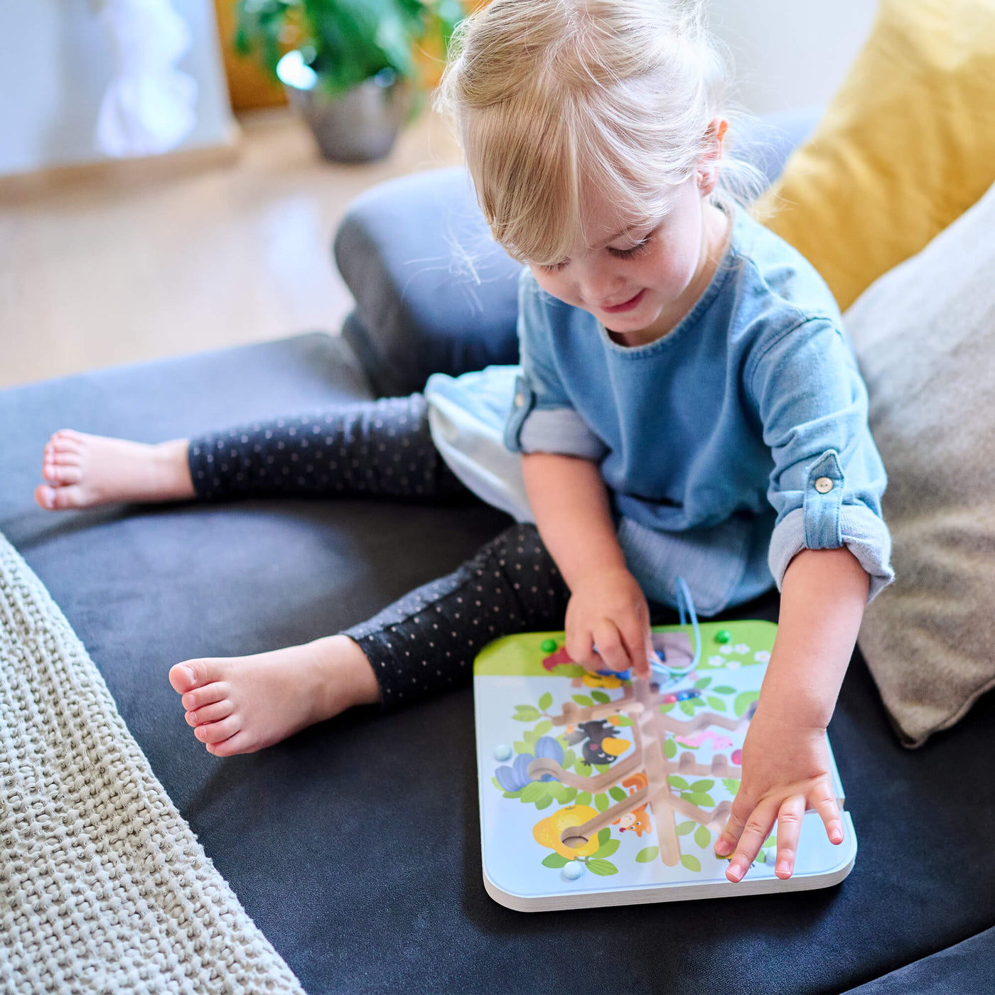 A young girl with blonde hair sits on a couch, playing with a colorful wooden puzzle called Orchard Maze Magnetic Sorting Game 