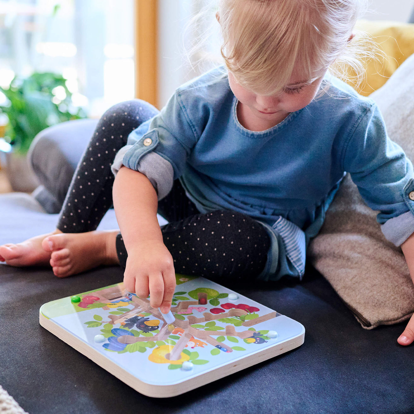 A young child with blonde hair plays on a sofa, focused on Orchard Maze Magnetic Sorting Game featuring trees fruit, and ravens.