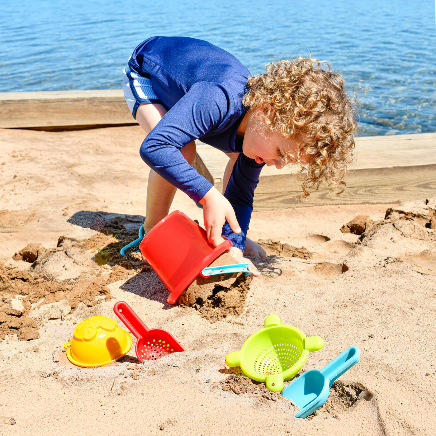 A young child with curly hair plays in the sand by a lake, using a red bucket with the HABA 5 Piece Basic Sand Toys Set  scattered nearby.